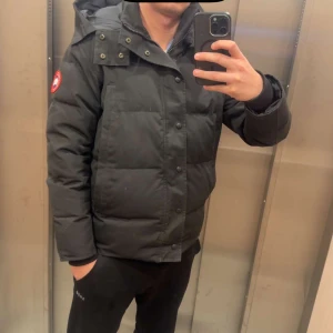  Canada Goose  - Säljer en svart dunjacka från Canada Goose med huva och ikonisk röd logga på ärmen. Jackan är quiltad och har både dragkedja och knappar framtill. Perfekt för kalla dagar och har en clean, modern look som funkar till streetwear.