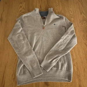 Grå merinoulltröja från Morris - Snygg grå långärmad tröja från Morris i 100% merinoull. Tröjan har half zip med läderdetalj på dragkedjan och en broderad blå logga på bröstet. Ribbstickade muddar och krage med blå insida ger en clean och stilren look. Tröjan är i storlek M men passar som S