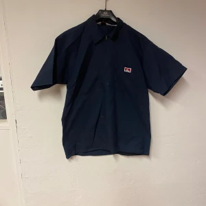 Mörkblå Ben Davis kortärmad skjorta - Mörkblå kortärmad skjorta från Ben Davis med klassisk krage, bröstficka och dragkedja framtill. Skjortan har en ikonisk Ben Davis-patch på bröstet och är tillverkad i en mix av bomull och polyester. Perfekt för en clean och avslappnad stil.