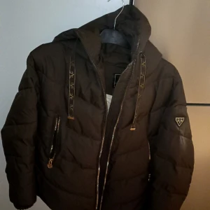 Brun pufferjacka från 19v69  Italia  - Snygg svart pufferjacka från V1969 Italia med huva och coola detaljer som logoband och emblem på ärmen. Jackan har dragkedja framtill och två fickor med dragkedja. Perfekt för dig som vill ha en varm och stilren look under kalla dagar. Tagit väl hand om den använt den max 3 ggr ÄKTA  