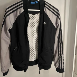 Svart och grå bomberjacka Adidas - Snygg svart och grå bomberjacka från Adidas med klassiska tre ränder längs ärmarna och Adidas-logga på bröstet. Jackan har dragkedja framtill, två fickor med dragkedja och ribbade muddar. Har en fläck på armen som visas på bilen. 