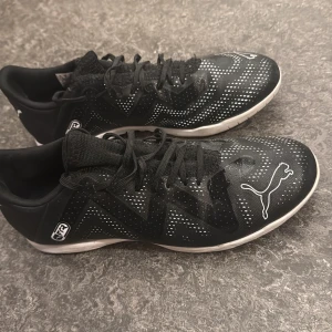 Svarta fotbollsskor/inomhusskor från Puma - Säljer ett par svarta fotbollsskor från Puma med vita detaljer och logga på tån. Skorna har snörning och en låg profil med platt sula, perfekt för inomhusspel. Ovandelen är i syntetmaterial med ett mönster av små vita prickar för extra stil.