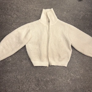 Vit stickad kofta med dragkedja - Mysig vit stickad kofta med hög krage och hel dragkedja framtill. Koftan har långärmad design och ribbad struktur som ger en stilren och enkel look. Perfekt för lager-på-lager under kyliga dagar.