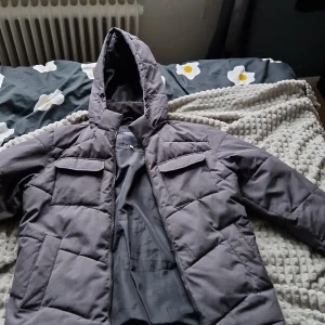 Grå pufferjacka från Produkt S - Säljer en grå pufferjacka från Produkt med huva och praktiska fickor framtill. Jackan har quiltad design och dragkedja hela vägen upp. Perfekt för kalla dagar och har en modern dansk stil. Passar dig som gillar enkel och clean look.