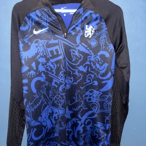 Nike Chelsea Dri-Fit långärmad tröja - Säljer en Chelsea långärmad träningströja från Nike med blått och svart grafiskt mönster, halv dragkedja och klubbens lejonlogga på bröstet. Tröjan är i slim fit och tillverkad i Dri-Fit polyester som andas. Perfekt för fotbollsträning eller chill. Skriv om ni har några frågor