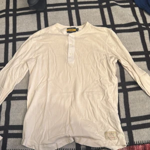 Vit långärmad tröja från Ralph Lauren - Vit långärmad tröja från Ralph Lauren Rugby med rund hals och två knappar framtill. Tröjan har en enkel, clean look och är gjord i mjuk bomull. Liten patch med Rugby-logga nedtill. Perfekt för dig som gillar klassisk och avslappnad stil.