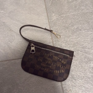 Brun kuvertväska - Snygg brun kuvertväska med klassiskt rutigt mönster och guldfärgad dragkedja. Väskan har en smidig handledsrem med metallspänne och tryckt logga i guld på framsidan. Perfekt storlek för det viktigaste och riktigt ikonisk design.