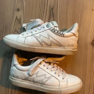 Zadig & Voltaire vita sneakers med blixt - Snygga vita sneakers från Zadig & Voltaire med coolt blixtmönster i silvernitar på sidorna och texten 'AMOUR' broderad vid hälen. Skorna har rund tå, snörning och är tillverkade i skinn. Perfekta för dig som vill sticka ut med detaljer.