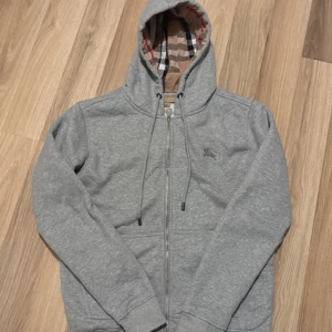 Grå Burberry hoodie med dragkedja - Snygg grå hoodie från Burberry med klassiskt rutigt foder i huvan. Tröjan har dragkedja framtill, snörning i huvan och ribbade muddar. Perfekt för dig som gillar stilrena plagg med en touch av lyx.