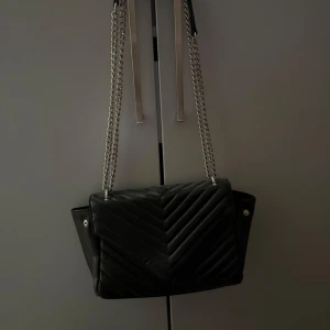 Svart quiltad axelväska från Gina Tricot - Snygg svart axelväska från Gina Tricot med quiltad framsida och silverfärgad kedja som axelrem. Väskan har ett stort fack med dragkedja inuti och stilren design med v-format mönster på locket. Perfekt för att lyfta din outfit.