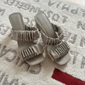 Beige sandaler med klack från H&M - Snygga beige sandaler från H&M med två breda, rynkade remmar och fyrkantig tå. Skorna har blockklack och öppen tå, vilket ger en trendig vibe. Tillverkade i syntetmaterial och perfekta för att lyfta din outfit.