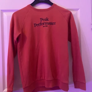 Röd Peak Performance sweatshirt - Säljer en röd sweatshirt från Peak Performance med svart logga på bröstet. Tröjan har rund hals, långa ärmar och ribbade muddar vid ärmslut och nederkant. Perfekt för dig som gillar en enkel och stilren look.