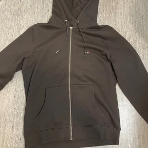 Svart hoodie från Tommy Hilfiger - Svart hoodie från Tommy Hilfiger med dragkedja framtill, huva med snörning och klassisk känguruficka. Liten Hilfiger-logga på bröstet och diskret röd-vit detalj på insidan. Perfekt för chill dagar och streetstyle. Bara testad inte använd. Skriv vid minsta fundering!