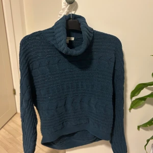 Mörkblå stickad polotröja (Derek Heart)  - Mysig mörkblå stickad polotröja från Stockh lm med grov kabelstickning och hög krage. Tröjan har långärmade ärmar och en loose passform, perfekt för kyliga dagar. Snygg att matcha med jeans eller kjol för en chill look.