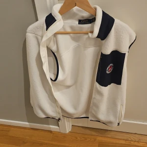 Vit fleeceväst från Polo Ralph Lauren - Säljer en vit fleeceväst från Polo Ralph Lauren med marinblå detaljer vid krage och bröstficka. Västen har dragkedja framtill och en snygg patch på bröstet. Perfekt för lager-på-lager och chill stil. Aldrig använda 
