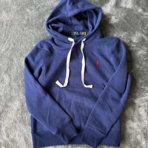 blå hoodie från Polo Ralph Lauren - Snygg marinblå hoodie från Polo Ralph Lauren med vit snörning och klassisk röd logobrodering på bröstet. Slim fit-modell med mjukt bomullsmaterial och stor huva. Perfekt för chill dagar och streetstyle. Känguruficka framtill för extra casual vibe.