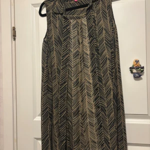 Beige och svart ärmlös långklänning - Säljer en ärmlös lång tunika i olivgrönt  och svart med ett coolt grafiskt mönster som påminner om pilar eller fjädrar. Tunikan har knappar fram och sprund i sidorna och är tillverkad i ett lätt, flowy material som känns luftigt och skönt.