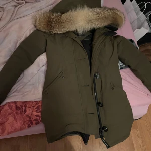 Canada Goose parkas med pälsluva - Säljer en mörkgrön Canada Goose parkas med stor fluffig päls på luvan. Jackan har två snedställda fickor, dragkedja och knappar framtill samt klassisk Canada Goose-märke på ärmen. Insidan är svart och jackan är fodrad för extra värme. Finns i Norrköping och hämta! Går med frakt också 