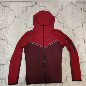 Röd Tech Fleece från Nike - Nike hoodie i två nyanser av rött med hel dragkedja och huva. Snyggt V-format mönster framtill och svart Nike-logga på bröstet. Långärmad med praktisk ficka med dragkedja på ena ärmen. Perfekt för chill eller träning.