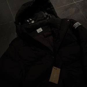 Svart pufferjacka från Burberry - Svart pufferjacka från Burberry med huva och klassiskt rutigt foder som går att ta av armarna för att omvandla till väst. Jackan har dragkedja, tryckknappar och en patch med 'Burberry London England' på ärmen. Snygg, varm och perfekt för kalla dagar. Stilsäker look med ikoniska Burberry-detaljer.