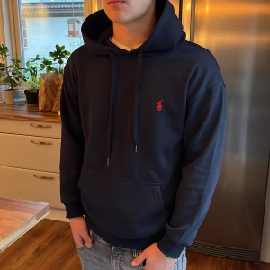 Marinblå hoodie från Polo Ralph Lauren - Snygg marinblå hoodie från Polo Ralph Lauren med röd broderad logga på bröstet. Tröjan har huva med snörning, stor magficka och ribbade muddar. Perfekt för dig som gillar klassisk stil med en sportig touch.