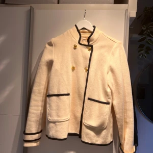 Beige rock från Holly & Whyte Lindex - Snygg beige rock med svarta detaljer från Holly & Whyte by Lindex. Jackan har guldknappar framtill, svarta kantband och två fickor. Ståkrage och långärmad modell, perfekt för dig som gillar klassisk stil med en modern twist.