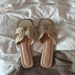 Beige sandaler med flätad detalj - Snygga sandaler i beige med flätad rem över foten och fyrkantig tå. Sulan är ljusrosa och har en platt design som gör dem sköna att gå i. Perfekta för varma dagar och har en trendig boho-vibe med sitt stickade material.