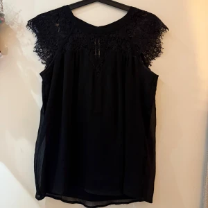 Svart blus med spetsdetaljer - Svart blus med vackra spetsdetaljer över axlar och bröst. Blusen är ärmlös och har en luftig, lätt passform med rund halsringning. Spetsen har ett blommigt mönster som ger en elegant touch. Perfekt att styla med jeans eller kjol för en cool look.