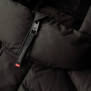 Svart pufferjacka från Superdry - Säljer en svart pufferjacka från Superdry med hög krage och huva. Jackan har synliga dragkedjor med Superdry-logga, två sidofickor och quiltad design. Perfekt för kalla dagar och har en clean, modern look som funkar till streetwear.