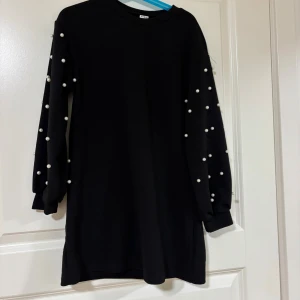 Svart klänning med pärldetaljer Shein - Svart kortklänning från Shein med långa ärmar dekorerade med vita pärlor. Klänningen har en rak passform och rund halsringning. Materialet känns mjukt och bekvämt, perfekt för dig som gillar stilrena plagg med en twist.