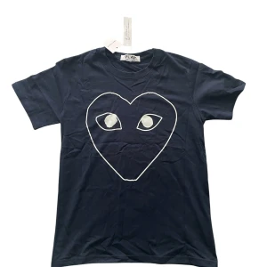 CDG t shirt - Snygg mörkblå t-shirt från CDG med det ikoniska hjärtmotivet med ögon i vitt på bröstet. Klassisk passform, rund hals och korta ärmar. 