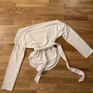 Vit ribbad omlottopp med knytband - Säljer en fräsch vit omlottopp med långa ärmar och snygga ribbade detaljer. Toppen har ett brett knytband i midjan som ger en cool och figurnära siluett. Perfekt för dig som gillar stilrena och trendiga plagg med lite extra edge.