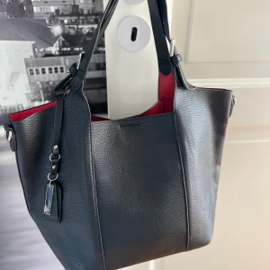 Svart axelväska med röd insida - Stilren svart axelväska i skinn med röd insida. Väskan har en rymlig form, praktiska innerfickor med dragkedja och silverfärgade detaljer. Perfekt för dig som vill ha plats för allt och samtidigt hålla looken clean och modern.