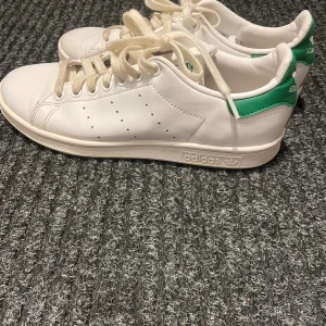 Adidas Stan Smith vita sneakers - Klassiska Adidas Stan Smith sneakers i vitt skinn med gröna detaljer på hälen och logga. Modellen har rund tå, platt sula och snörning framtill. Perfekt för dig som gillar tidlös och clean stil. Ikonisk design med perforerade ränder på sidorna.