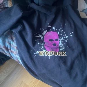 Svart hoodie med EPADUNK tryck - Svart hoodie med stor huva och fett EPADUNK-tryck på bröstet. Trycket har en rosa rånarluva och coola vita färgstänk runt motivet. Hoodien har ribbade muddar och klassisk känguruficka framtill. Perfekt för dig som gillar street och vill sticka ut.