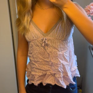 Ljusrosa prickig linnetopp med volanger - Supersöt ljusrosa linnetopp med små prickar (blommor) och volangdetaljer längst ner. Toppen har v-ringning med spetskant och en liten rosett framtill. Den är ärmlös och har en luftig, feminin passform som är perfekt för varma dagar. Jätte söt men jag har knappt användt den💕💕