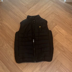 Svart dunväst från Lyle & Scott - Svart dunväst från Lyle & Scott. Västen har ståkrage och fickor vid sidorna samt inneficka. Juniorstorlek str 164-170