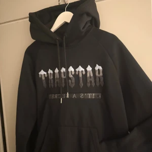 Svart hoodie från Trapstar - Säljer en svart hoodie från Trapstar med silverfärgat tryck på bröstet och snörning vid huvan. Hoodien har en stor magficka och coolt grafiskt tryck som ger streetkänsla. Perfekt för dig som gillar urban stil.