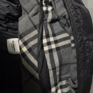 Svart dunväst från Burberry - Snygg svart dunväst från Burberry med klassiskt rutigt foder i grått, vitt och svart. Västen har quiltad design, dragkedja framtill och en Burberry London England patch på bröstet. Perfekt för att addera lite lyx till din look.