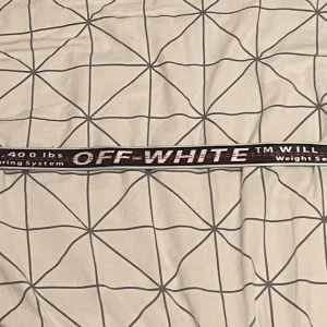 Svart Off-White bälte med text - Säljer ett svart bälte från Off-White med vit och röd text längs hela bandet. Bältet har en metallring som spänne och är gjort i syntetmaterial. Den ikoniska Off-White-loggan syns tydligt och bältet har en streetwear-vibe med industriell känsla.