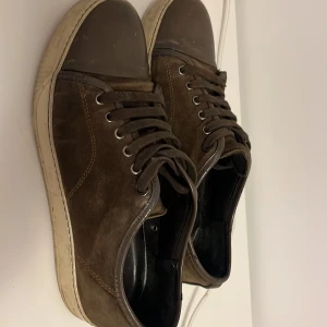 Mörk grön sneakers från Lanvin i mocka - Snygga bruna sneakers från Lanvin med rund tå och platt vit sula. Skorna har snörning och är tillverkade i mjuk mocka med detaljer i skinn. Klassisk design med kontrastsömmar och Lanvin-logga under sulan. Perfekta för dig som gillar stilrena och exklusiva sneakers.