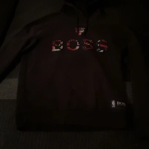 Svart Chicago Bulls hoodie NBA - Svart hoodie från BOSS x NBA med Chicago Bulls-tryck i rött och vitt framtill. Klassisk huva med snörning och lång ärm. Perfekt för dig som gillar basket och streetwear. Bulls-logga och NBA-märke nertill.