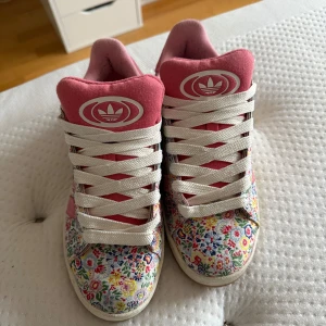Adidas sneakers blommiga rosa 39 - Färgglada Adidas sneakers i storlek 39 med blommigt mönster på tygdelen och rosa detaljer på plös och häl. Skorna har vita snören och gummisula i ljusbrun färg. Perfekta för dig som vill sticka ut med en unik och lekfull stil.