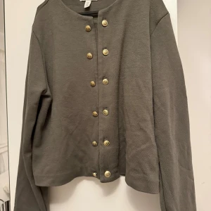Grå cardigan jacka från H&M med guldknappar - Snygg grå cardigan jacka från H&M i storlek M. Jackan har rund halsringning och är dekorerad med dubbla rader guldknappar framtill. Materialet är mjukt och känns som bomull, perfekt för lager-på-lager. Långärmad och med en loose passform.