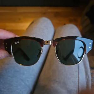 Ray-Ban Mega Clubmaster solglasögon - Snygga Ray-Ban Mega Clubmaster solglasögon med klassisk brun och svart båge i retrostil, detaljer i guld och gröna linser. Tillverkade i Italien, modell RB0316-S. Perfekt för dig som vill ha en ikonisk look med cool vibe.