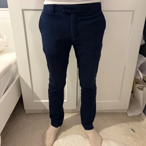 Mörkblå chinos Pier One, strl 46 - Snygga mörkblå chinos från Pier One i storlek 46. Byxorna har smal passform, bälteshällor, framfickor och bakfickor med knappar. Perfekt för dig som gillar stilrena och klassiska plagg med modern touch.