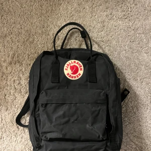Svart Fjällräven Kånken ryggsäck - Klassisk svart Fjällräven Kånken ryggsäck med ikonisk rund logotyp framtill. Ryggsäcken har ett stort huvudfack med dragkedja, en mindre ficka framtill och två sidofickor. Tillverkad i slitstark syntet och har både handtag och justerbara axelremmar.