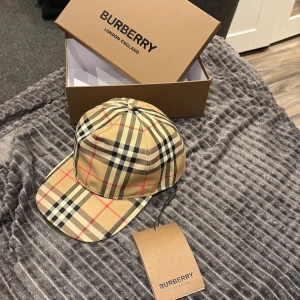 Burberry rutig keps i beige M - Säljer en klassisk keps från Burberry i ikoniskt beige rutigt mönster med svarta, vita och röda detaljer. Kepsen har justerbar baksida och tydlig Burberry-logga. Tillverkad i bomull och polyester, perfekt för dig som vill ha en stilren accessoar.