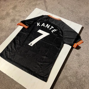 Chelsea Kanté #7 svart Nike fotbollströja - Chelsea FC bortatröja med Kanté #7 på ryggen. Svart med subtilt geometriskt mönster, vita tryck och orange-vita detaljer på krage och ärmslut. Tillverkad i lätt, ventilerande material. Nike-logga och Yokohama Tyres sponsortryck på bröstet.