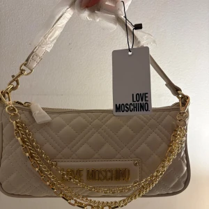 Beige quiltad axelväska Love Moschino - Snygg beige axelväska från Love Moschino med quiltat mönster och guldfärgade kedjedetaljer. Väskan har en stilren form, dragkedja upptill och ett stort M broderat på baksidan. Materialet är syntet och den har en exklusiv vibe med logotyp framtill.